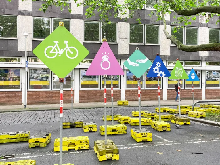 Mehrere bunte Schilder auf einem Platz. Im Hintergrund ein Gebäude.